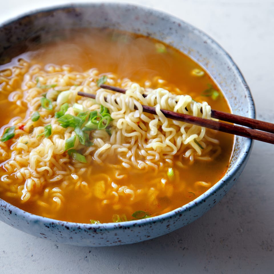 Bowl of spicy ramen with curly noodles and sliced green onions, served with chopsticks in a speckled ceramic bowl.