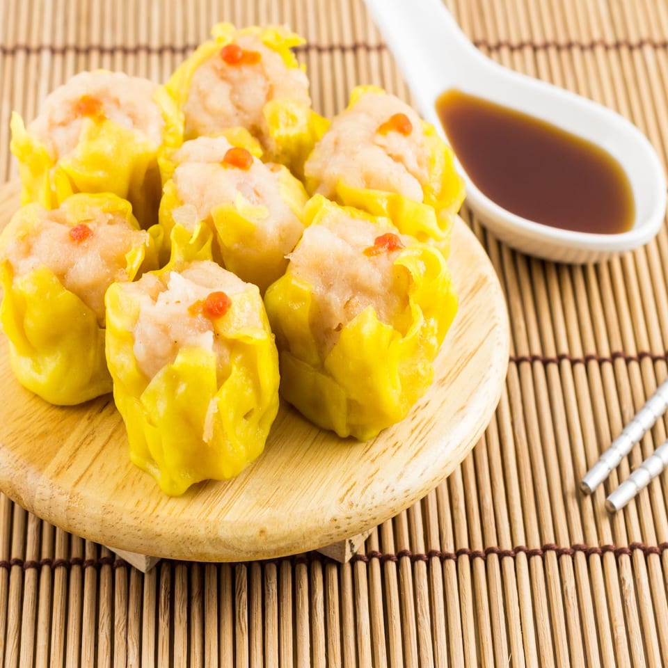 Steamed yellow siu mai dumplings topped with orange roe on a wooden plate, served with soy dipping sauce and chopsticks.